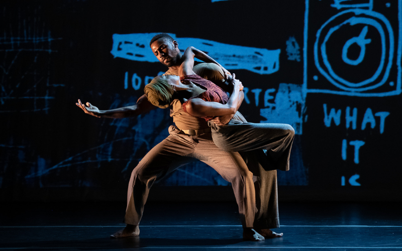 Two dancers performing on a stage with a dark background containing blue drawings