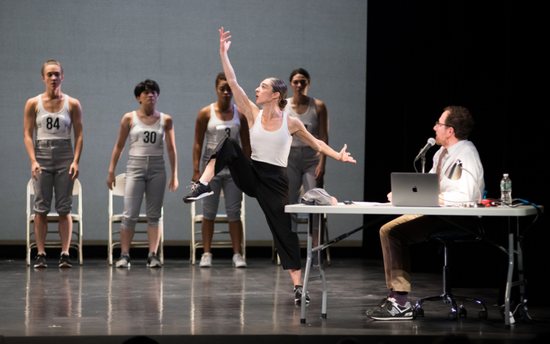 A dancer in front of a line of performers waiting to dance and a person on a table watching on