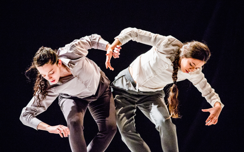 two dancers wearing similar clothing pose as mirror images on stage