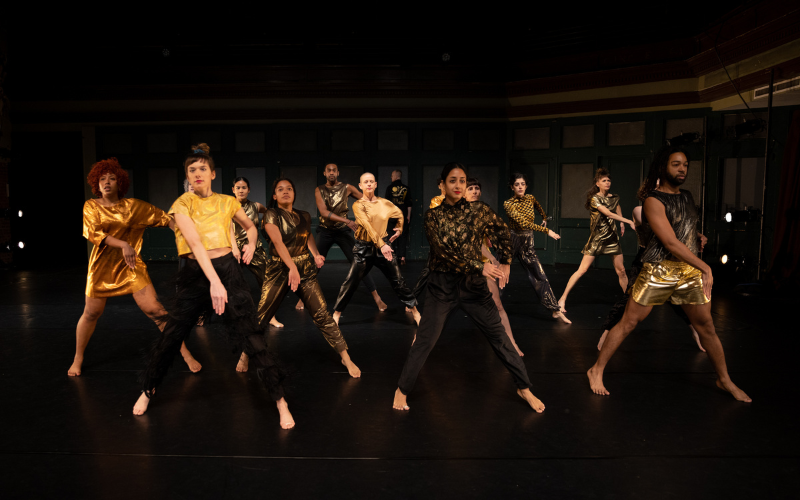 Many dancers wearing black and gold stand leaning to their right with their right arm extended downward across their bodies.