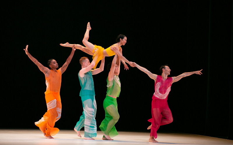4 dancers in colorful costume lift a 5th above their heads on stage.